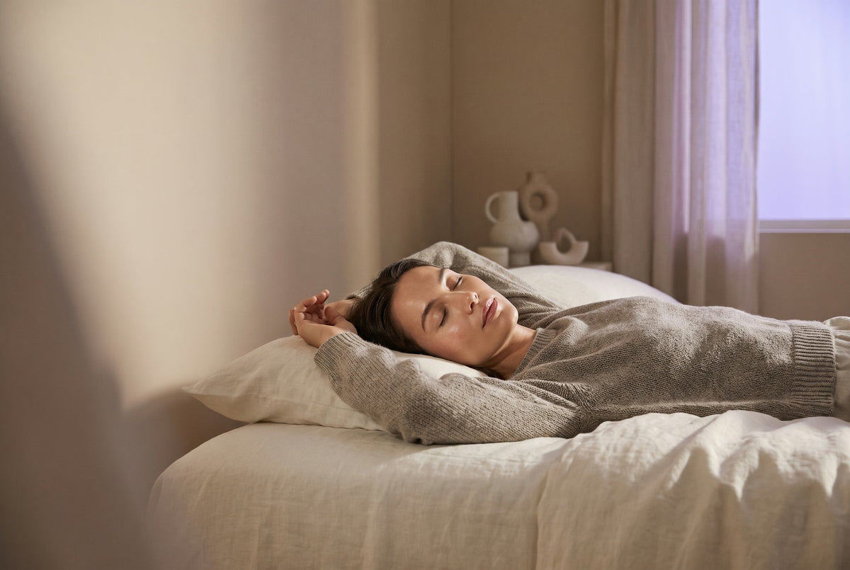 Model resting in bed wearing an eye mask in soft morning light.