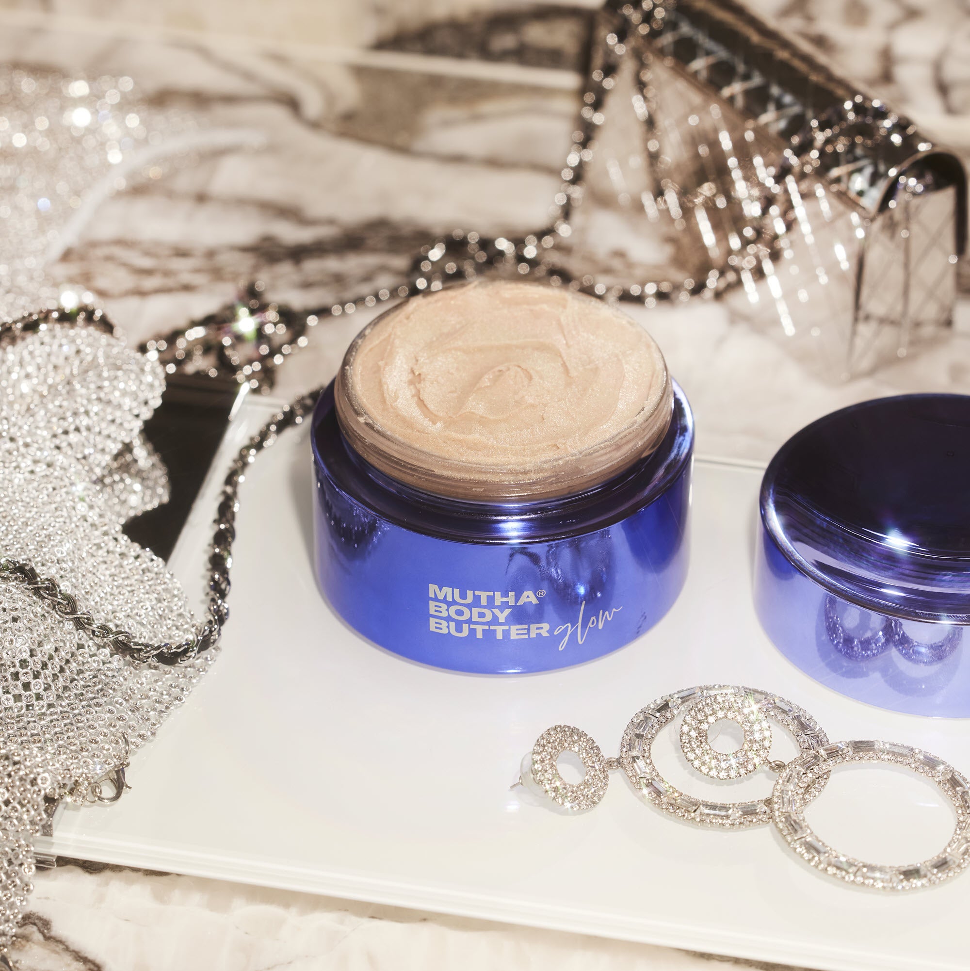 Stylish image of the Body Butter Glow opened and on a marble counter top with sparkly clothing, a purse, and jewelry