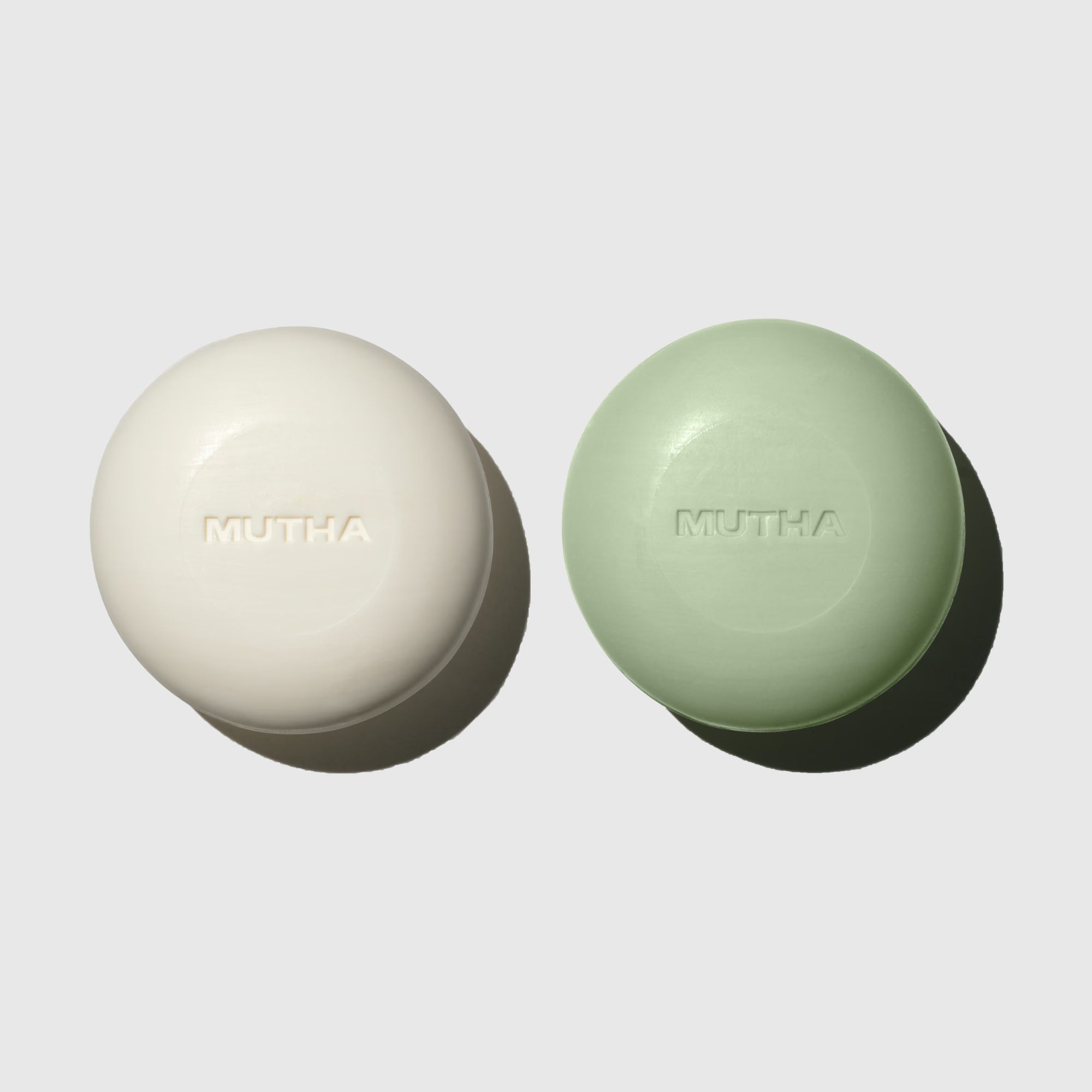 MUTHA Exfoliating Body Bars in Dirty Citrus and Detox Greens side by side on a soft grey background, each embossed with the MUTHA logo