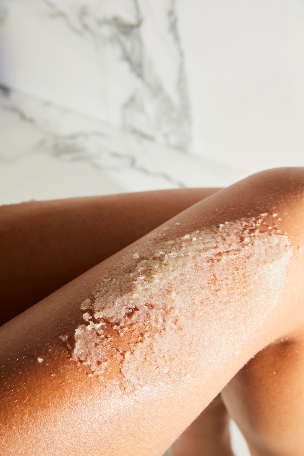 Close-up of a model's leg with MUTHA Body Scrub sugar textured scrub on a marble surface