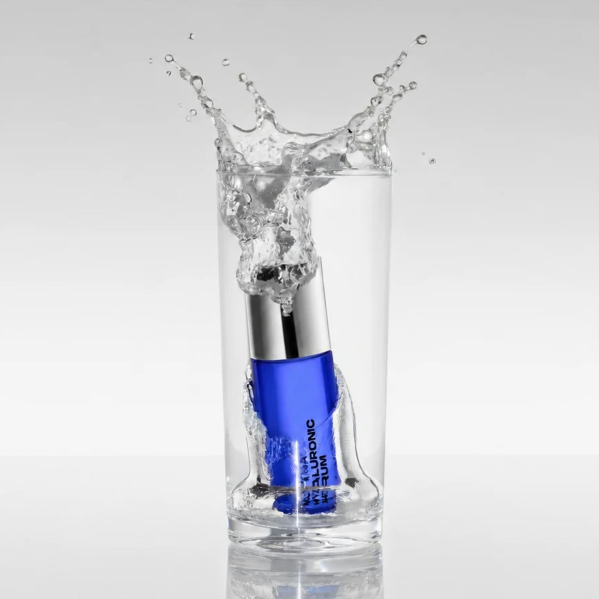 The cobalt blue MUTHA Hyaluronic Serum bottle dropping into a tall glass of water, sending a splash over the rim