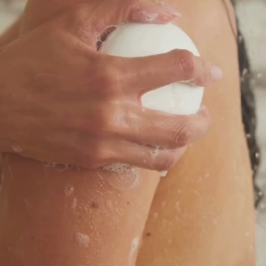 A model lathers the white Dirty Citrus body bar over wet skin in the shower, building a rich foam