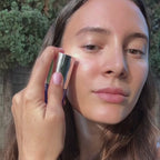 A model applies MUTHA Hyaluronic Serum to her face in natural daylight, pressing it under the eyes and across the cheeks