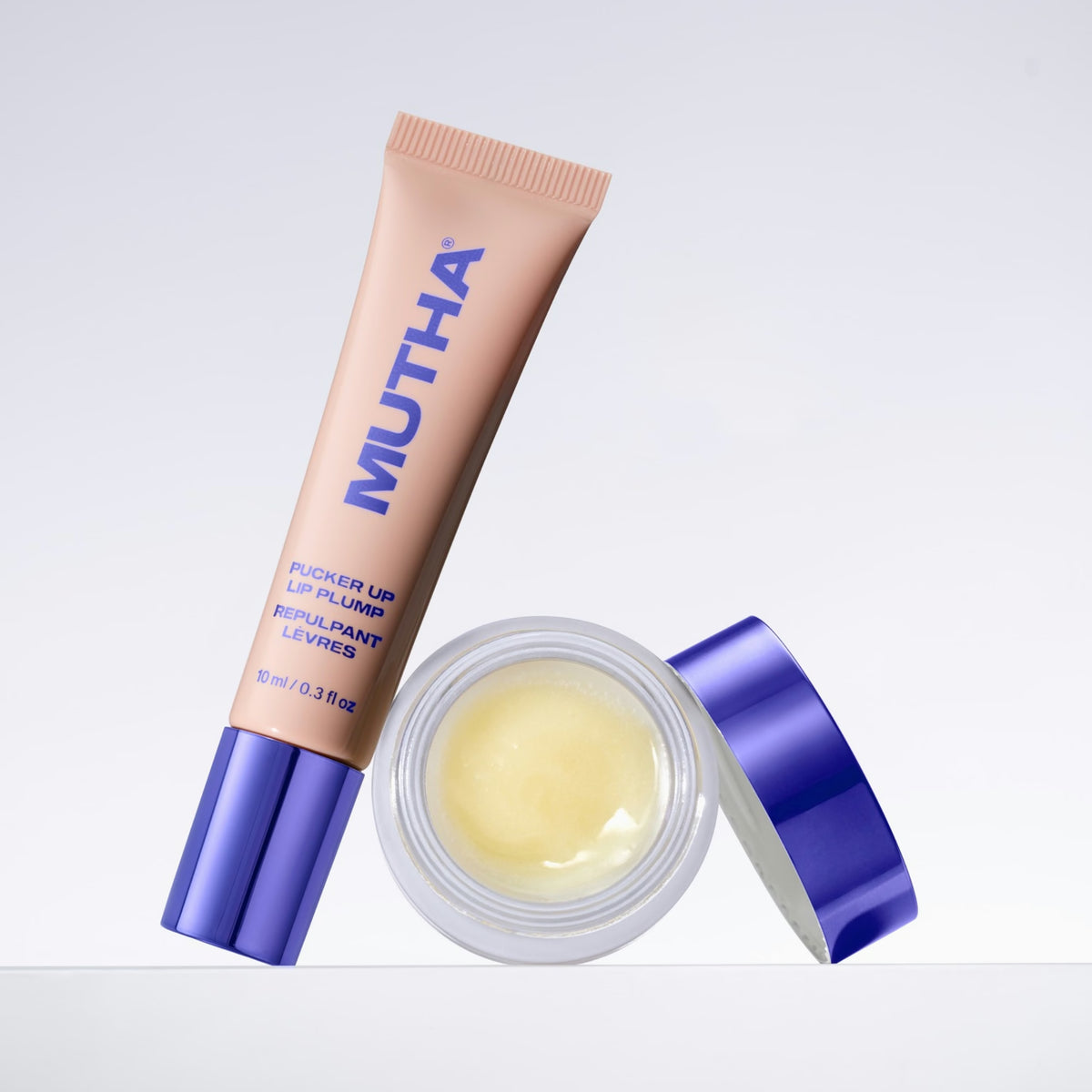 MUTHA Pucker Up Lip Plump tube leaning against an open jar of Pucker Lip Butter, the balm visible inside and the purple lid propped alongside
